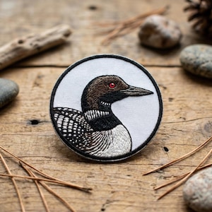 May include: Embroidered patch featuring a detailed illustration of a loon. The patch has a white background with a black border. The loon has brown and black feathers, a red eye, and a long, pointed beak. The patch is circular.