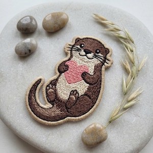 May include: Embroidered patch featuring a brown otter holding a pink heart. The otter has a cream-colored belly and is surrounded by a tan border. The patch is on a white surface with small stones and a sprig of dried grass.