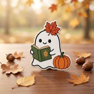 May include: An embroidered patch featuring a white ghost with a smiling face, holding an open green book with a gold leaf design. A vibrant orange pumpkin sits beside the ghost, and a single red maple leaf rests on top of its head.
