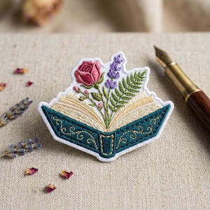 May include: Embroidered patch in the shape of an open book, featuring a pink rose, purple lavender, and green fern. The book's cover is teal with gold detailing, and the pages are beige. A decorative item for book lovers.
