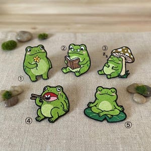 May include: A collection of five embroidered patches featuring cartoon frogs in various poses. One frog holds a flower, another reads a book, and a third sleeps under a mushroom. The other two frogs are eating and meditating.