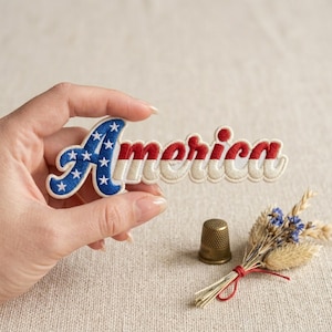 May include: Embroidered patch with the word "America" in red, white, and blue. The blue section features white stars. A small bouquet of dried flowers and a brass thimble are also visible.