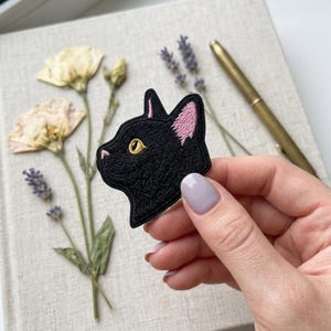 May include: Black cat patch with pink ear details and a gold eye. The embroidered patch is in the shape of a cat's head in profile. The patch is held in front of a notebook with dried flowers and a pen.