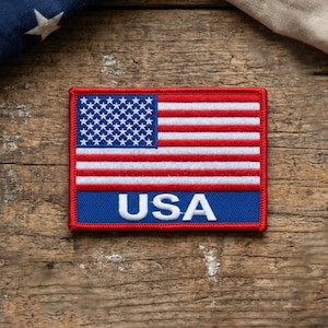May include: Rectangular patch featuring the American flag design. The top section displays a blue field with white stars, while the lower section has red and white stripes. The word "USA" is in white letters on a blue background.