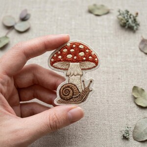 May include: Embroidered patch featuring a red mushroom with white spots and a snail. The mushroom has a beige stem and a brown snail is below it. The patch is held in a hand, with a neutral background.