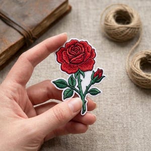 May include: An embroidered fabric patch featuring a vibrant red rose bloom with a smaller red rosebud, green leaves, and a green stem. The design is outlined in white thread and set against a white background, showcasing detailed stitching throughout.