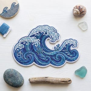 May include: An embroidered patch featuring a stylized ocean wave in shades of blue and white. The design includes a small sea turtle, a tiny anchor, and a school of fish integrated into the swirling water patterns with a white border edge.