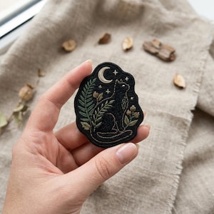 May include: Black embroidered patch featuring a cat, moon, stars, and botanical elements. The patch has a dark background with detailed stitching in green, gold, and white. The cat is sitting and looking up at the moon.