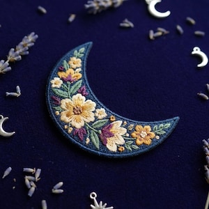 May include: Embroidered crescent moon patch with a floral design. The patch features yellow, purple, and green flowers on a navy blue background. Small silver charms and lavender sprigs surround the patch.
