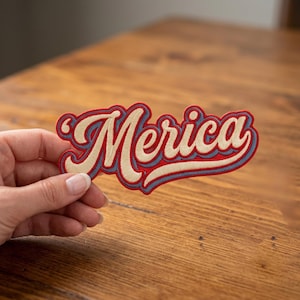 May include: Embroidered patch with the word "Merica" in a retro script font. The patch features a red border, a light blue shadow, and the word in a cream color. The patch is held in a hand.