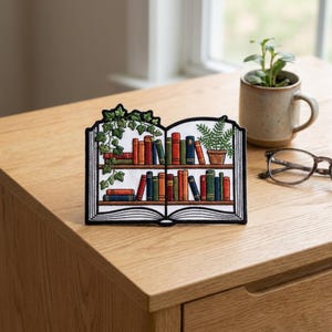 May include: Embroidered patch in the shape of an open book with two shelves of colorful books. Green vines and a potted fern decorate the top. The patch is on a wooden surface, with a mug and glasses in the background.