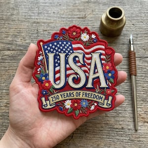 May include: Embroidered patch featuring the American flag and the word "USA" in white, surrounded by red, white, and blue flowers. The patch reads "250 YEARS OF FREEDOM" on a banner. A patriotic emblem.