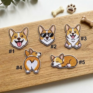 May include: Embroidered patches featuring corgi dogs in various poses, including a face with sunglasses, a sitting corgi with its tongue out, and a corgi from behind. The patches are in shades of brown, white, and black.