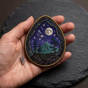 May include: Embroidered patch featuring a night landscape with a mountain, trees, stars, and a full moon. The patch has a dark blue and green color scheme, with a detailed design and a dark border.