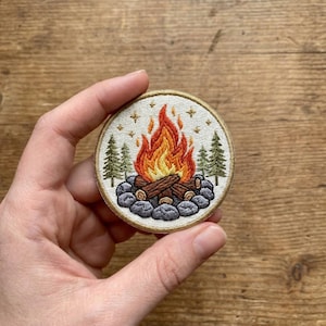 May include: Round embroidered patch featuring a campfire scene. The design includes a vibrant orange and yellow flame, brown logs, gray stones, green pine trees, and small gold stars. The patch has a cream-colored background and a gold border.