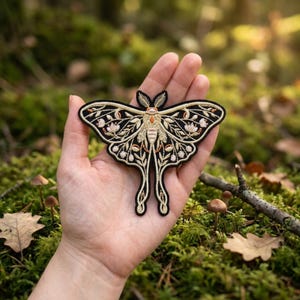 May include: Embroidered moth patch with a black outline, gold body, and floral details. The patch features a detailed design of a moth with intricate patterns and delicate stitching. This decorative patch can be applied to clothing or accessories.