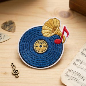 May include: Embroidered patch featuring a blue vinyl record with a gold gramophone and a red musical note. The patch has a white border. Musical theme.