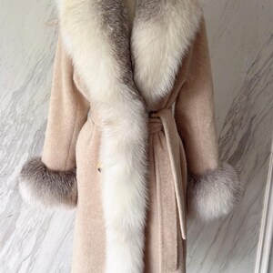 May include: A long, beige coat with a luxurious white and grey fur collar and cuffs. The coat features a matching belt and is displayed on a mannequin. A stylish outerwear garment.
