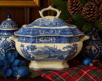 Op de afbeelding: Een grote, crèmekleurige terrine met een blauw-wit landschapsontwerp. De terrine heeft een deksel met een handvat en zijhandvatten. Het wordt weergegeven op een rood-groene geruite doek, met kerstversieringen op de achtergrond.