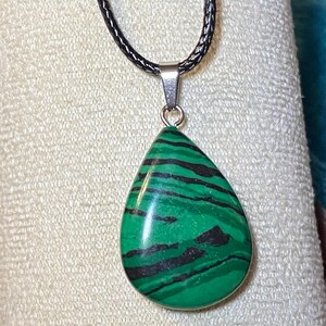 May include: A teardrop-shaped pendant necklace featuring a polished green stone with black stripes. The stone is suspended from a silver-toned bail and a black cord necklace. The pendant is approximately 1.5 inches long.