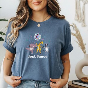 May include: A blue t-shirt featuring a whimsical design with a disco ball and dancing animal characters: a hippo, giraffe, and zebra. The words "Just Dance" are printed below the illustration. The shirt has short sleeves.