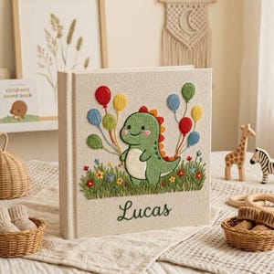 Może przedstawiać: Beżowa książka dla dziecka z haftowanym zielonym dinozaurem trzymającym kolorowe balony. Imię "Lucas" jest wyhaftowane poniżej. Książka stoi na białej powierzchni.
