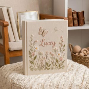 May include: A beige fabric-covered book with embroidered floral and butterfly details. The word "Luccy" is embroidered in pink script. The book is standing upright on a cream-colored knitted blanket.