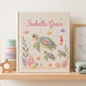 May include: A square book with a cream-colored fabric cover featuring embroidered sea life, including three sea turtles, a seahorse, fish, coral, and seashells. The name Isabella Grace is embroidered in pink script at the top center.