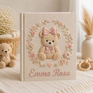 May include: A beige fabric-covered photo album featuring an embroidered design of a teddy bear with a pink bow, surrounded by a floral wreath. Below the bear, the name Emma Rosa is embroidered in pink script, with a small pink heart centered above it.