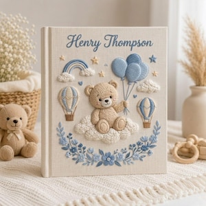 May include: A beige fabric-covered book featuring embroidered designs of a teddy bear sitting on a cloud, blue balloons, hot air balloons, a rainbow, and flowers. The text Henry Thompson is embroidered in blue script at the top of the cover.