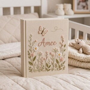 May include: A beige fabric-covered book with embroidered floral and butterfly details. The name "Amee" is stitched in pink script. The design includes daisies, other flowers, and green leaves, creating a delicate, handcrafted look.