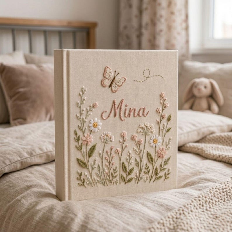 May include: A beige fabric-covered book with embroidered floral and butterfly details. The name "Mina" is embroidered in pink script. The cover features a butterfly and various flowers, creating a delicate, handcrafted appearance. The book is resting on a bed.