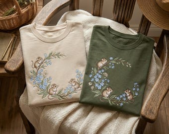 May include: Two crewneck t-shirts, one beige and one olive green, each featuring an embroidered circular wreath design. The embroidery depicts three small brown mice nestled among light blue flowers and green leafy vines.