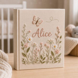May include: A beige fabric-covered book with embroidered floral and butterfly details. The name "Alice" is stitched in cursive. The cover features a butterfly and various flowers in pink, white, and green, creating a delicate, handcrafted aesthetic.