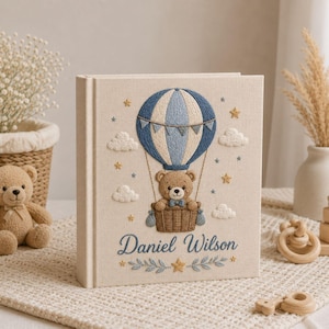 May include: A cream-colored fabric-covered photo album featuring an embroidered design of a brown teddy bear in a blue and white hot air balloon. The embroidery includes clouds, stars, and the name Daniel Wilson in blue script with leaf accents.