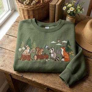 May include: An olive green sweatshirt with an embroidered woodland scene. The design features a rabbit, squirrel, raccoon, and fox, with trees, flowers, and a butterfly. The sweatshirt is folded on a wooden table.