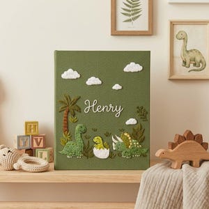 Może przedstawiać: Zielona okładka książki z haftowanymi dinozaurami, chmurami i palmą. Imię "Henry" jest napisane kursywą. Drewniane zabawki dinozaurów i oprawione dzieła sztuki znajdują się w tle.