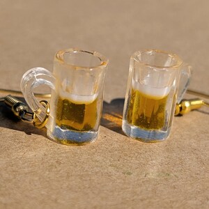 Könnte beinhalten: Ein Paar ausgefallene Ohrringe mit Miniatur-Bierkrügen. Jeder klare Glaskrug ist mit einer goldenen Flüssigkeit gefüllt, die Bier ähnelt, und hat eine weiße Schaumkrone. Die Ohrringe haben goldfarbene Ohrhaken.