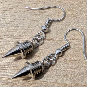 May include: A pair of silver-colored earrings featuring a spike design. Each earring has a pointed cone shape with a threaded base, connected to a hook and chain. The earrings are made of a shiny, metallic material.