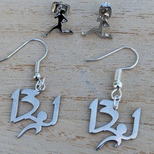May include: A silver jewellery set comprising two pairs of earrings. One pair are drop earrings with a stylized number 13 and a figure. The other pair are stud earrings with a running figure.