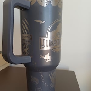 May include: A dark gray insulated tumbler with a large handle and a clear lid. The tumbler features etched designs of dinosaurs, foliage, and the word "JURASSIC". A warning label is also visible.