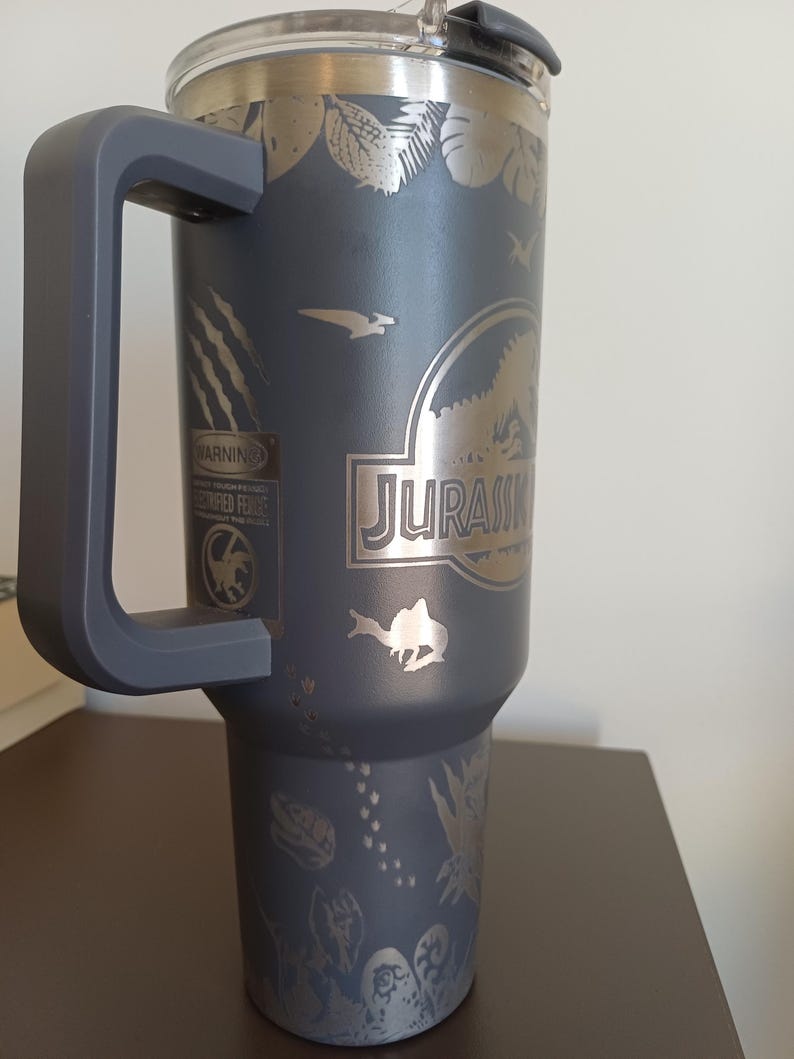 May include: A dark gray insulated tumbler with a large handle and a clear lid. The tumbler features etched designs of dinosaurs, foliage, and the word "JURASSIC". A warning label is also visible.
