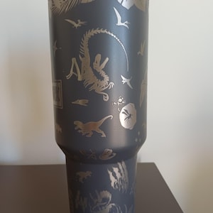 May include: A dark gray stainless steel tumbler with a silver dinosaur theme. The tumbler features etched images of various dinosaurs, pterodactyls, and tropical foliage. The word "PARK" is visible on the side.