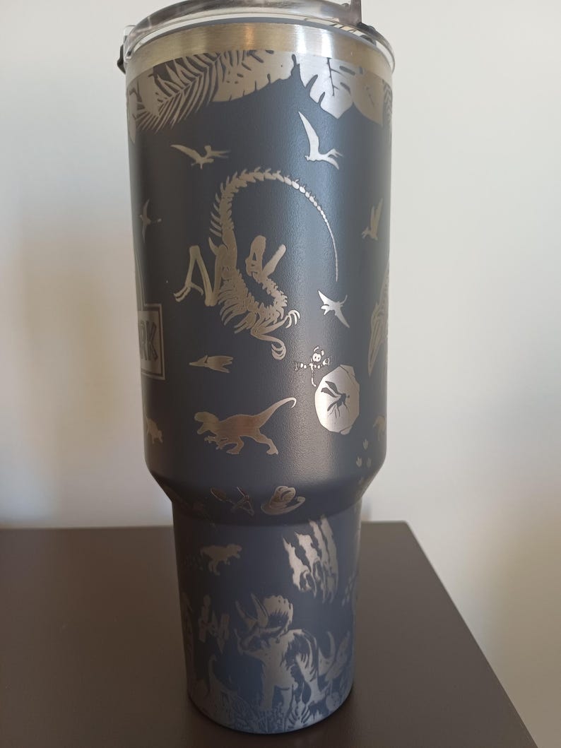 May include: A dark gray stainless steel tumbler with a silver dinosaur theme. The tumbler features etched images of various dinosaurs, pterodactyls, and tropical foliage. The word "PARK" is visible on the side.