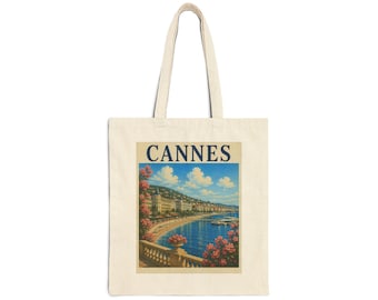 May include: A beige canvas tote bag featuring a rectangular print of a coastal scene in Cannes. The image displays a blue sea, a sandy beach, buildings along the shore, and pink flowers in the foreground. The word CANNES is printed in blue.