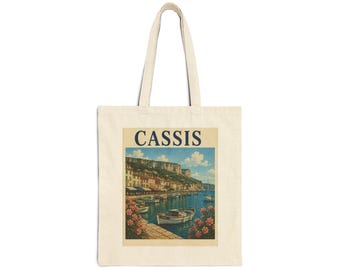May include: A beige canvas tote bag featuring a rectangular print of a coastal harbor scene in Cassis. The illustration depicts boats on blue water, colorful buildings along the shore, cliffs in the background, and pink flowers in the foreground.