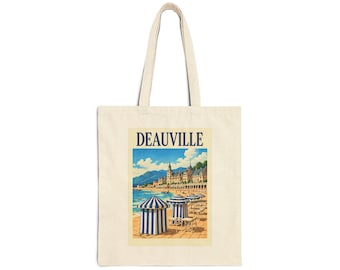 May include: A beige canvas tote bag featuring a rectangular graphic print of a beach scene in Deauville. The illustration depicts blue and white striped beach tents on a sandy shore with a coastal town and mountains in the background.