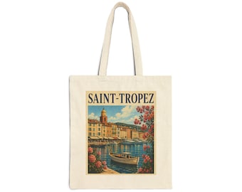 May include: A beige canvas tote bag featuring a rectangular graphic print of a Saint-Tropez harbor scene. The illustration depicts a small boat on blue water, coastal buildings, and pink flowers, with the text SAINT-TROPEZ printed in dark blue.