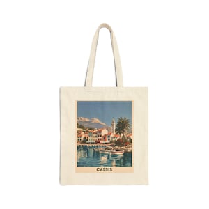 May include: A beige canvas tote bag featuring a rectangular print of a coastal scene with boats on the water, colorful buildings, a palm tree, and a mountain in the background. The word CASSIS is printed in black capital letters below the image.