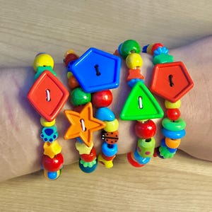 May include: Brightly coloured beaded bracelets with large, shaped buttons. The bracelets feature red, orange, blue, and green buttons in diamond, star, pentagon, and triangle shapes. The beads are various colours and sizes.